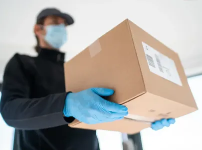 A person wearing a mask and gloves holds a box. Appears to be delivering the box.